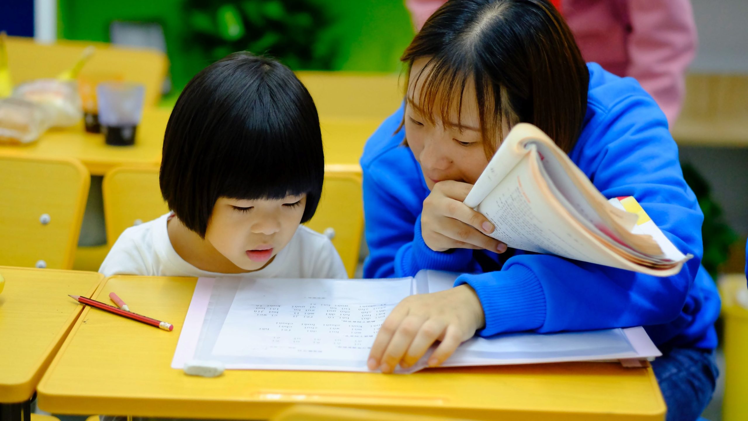 Woman helps child at a desk with academic work, presumably a reading or math assignment.